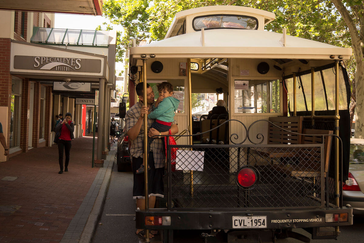 A man holding a toddler in a green sweater hangs onto the pole at the back of Subiaco's old fashioned, open tram