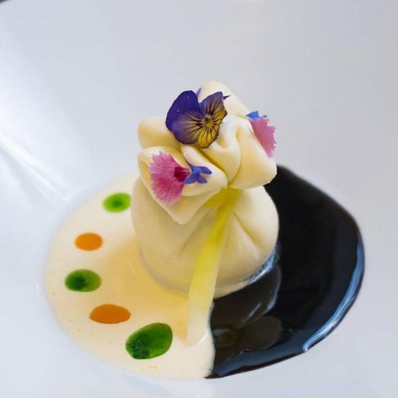 An ornate dumpling with edible flowers arranged on top of two sauces one dark one light