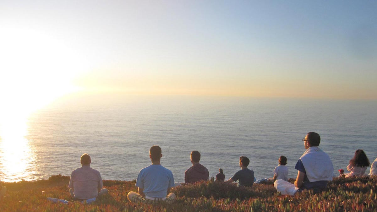 a group of people all sitting and meditating separately on a hill overlooking the water as the sun sets